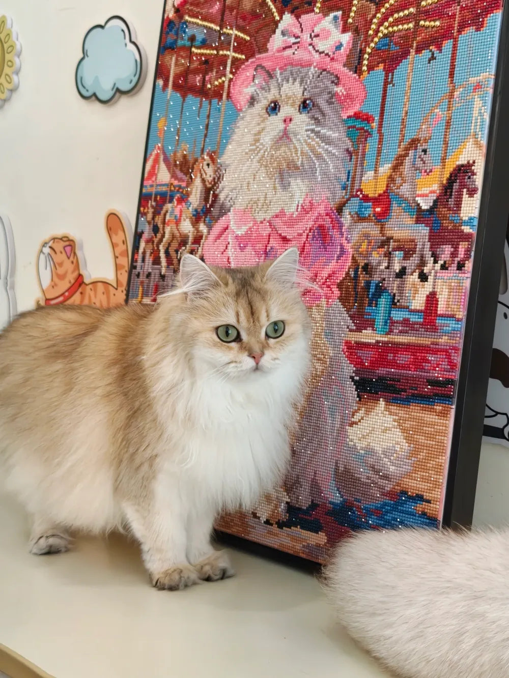 A fluffy golden cat poses beside a framed kitten diamond painting showing a dressed-up cat in a colorful carnival scene - FEELOOK Art