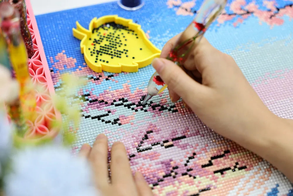 Hands placing colorful drills on a 5D diamond painting canvas with a yellow tray and diamond painting pen - FEELOOK Art